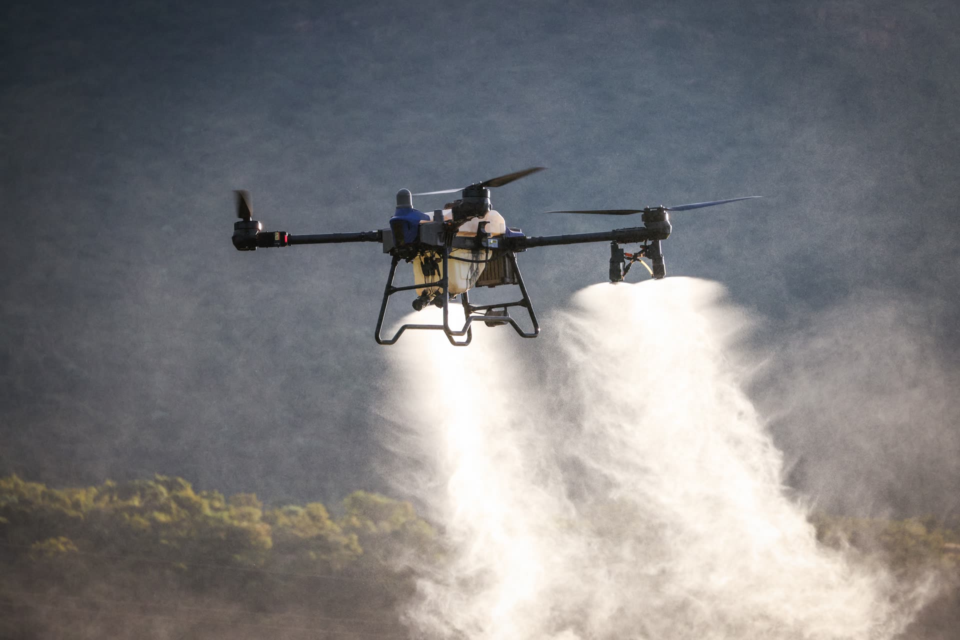 An agricultural drone spraying a field, backlit against the sky